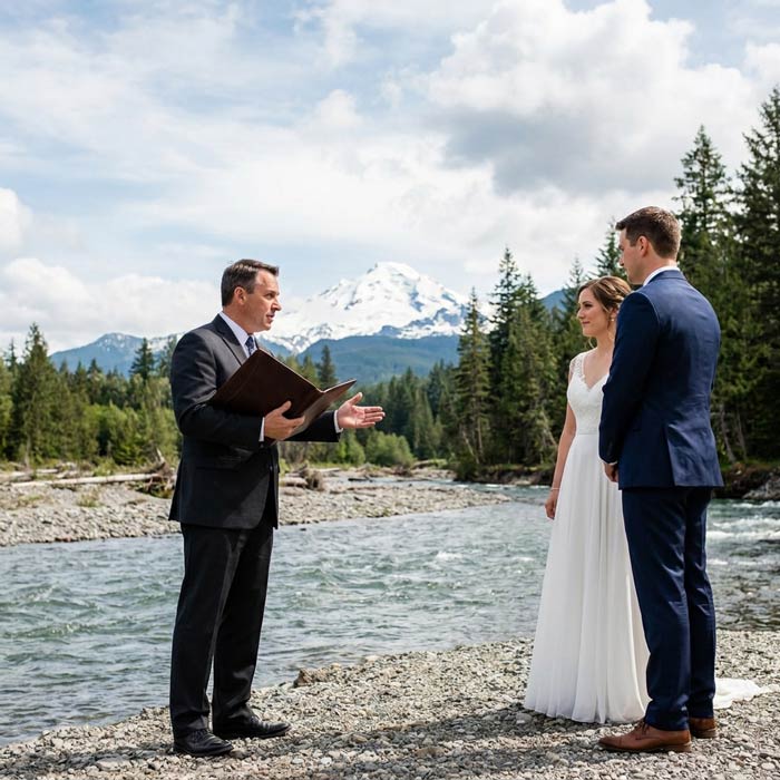 An officiant speaks to a couple during a mountain ceremony — photo reveals colour on hover